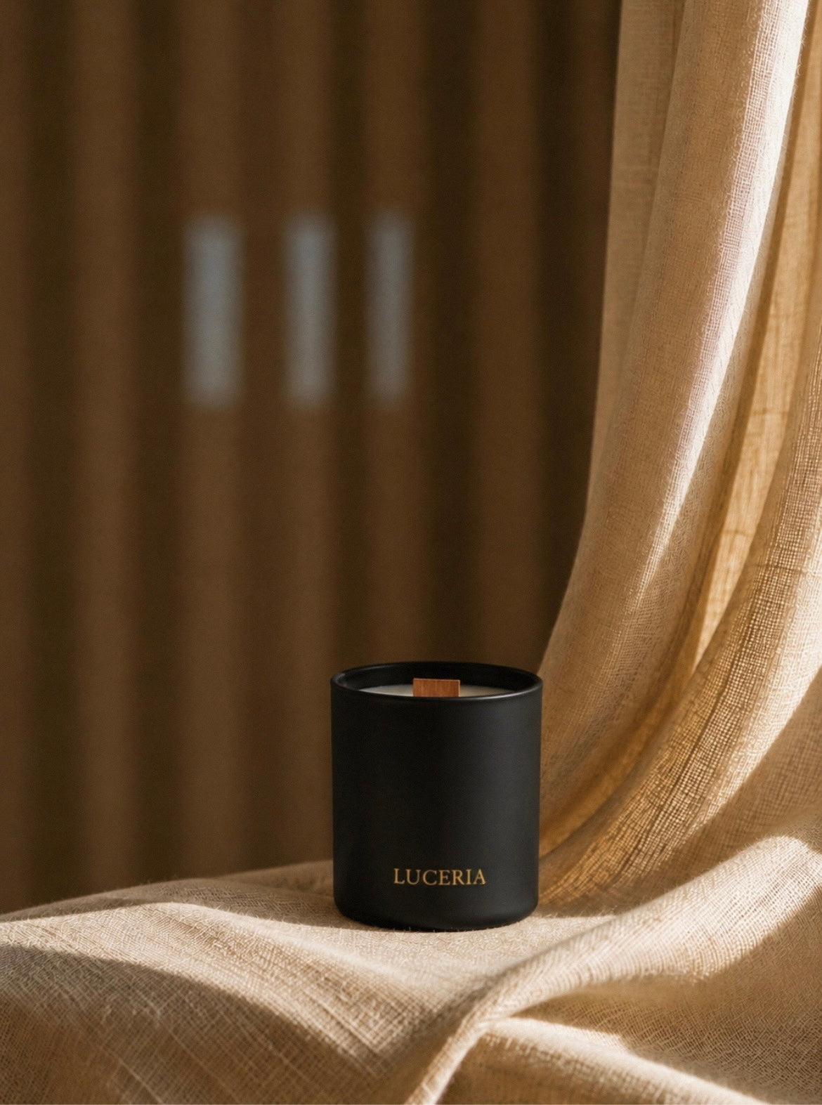Luceria matte black wood wick candle displayed on soft linen fabric beside sheer curtains in warm natural light.