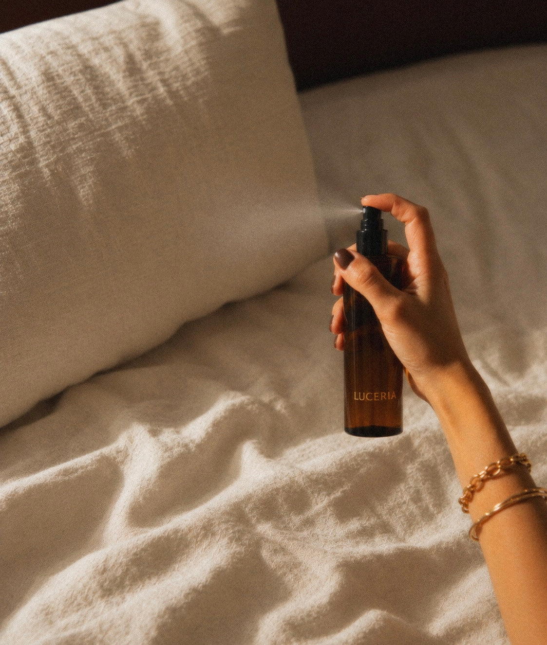Woman holding black cap amber spray bottle with LUCERIA branding misting the bed