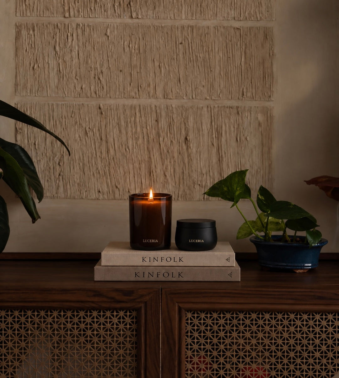 A lit amber glass candle and a small black jar labeled LUCERIA are placed on stacked KINFOLK books on a wooden cabinet, with potted plants on both sides against a textured beige wall.