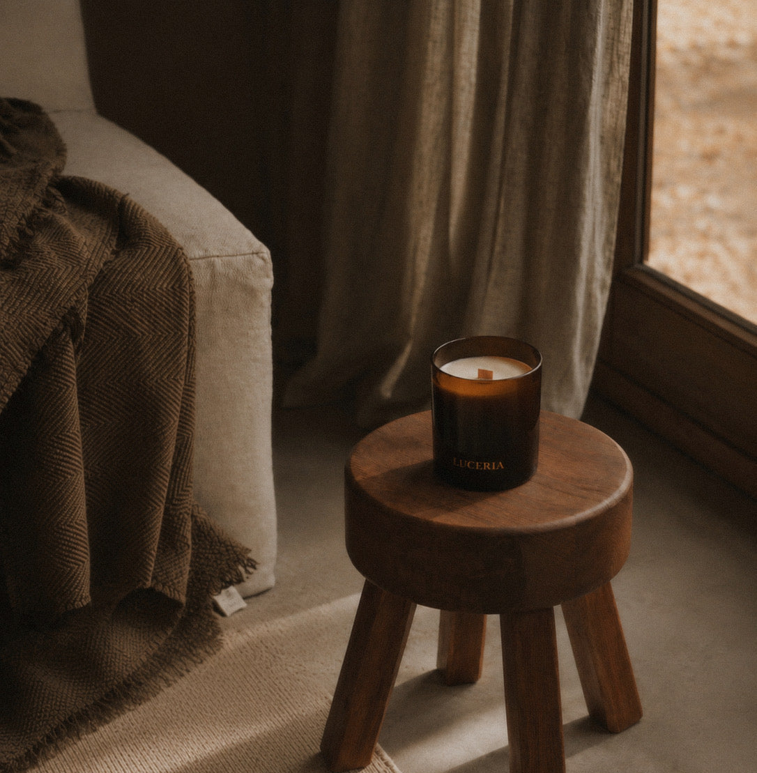 A Luceria candle on a wooden stool in a cozy room with a couch and curtains.