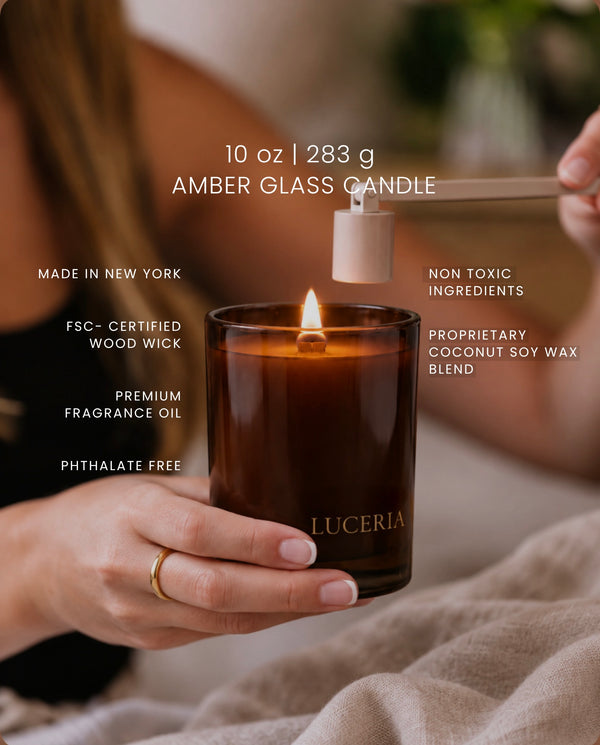 An amber glass candle held in a hand, with a wick being lit from above using a wooden tool. White text overlays highlight product features and ingredients, including '10 oz | 283 g AMBER GLASS CANDLE' and 'LUCERIA' printed on the jar.