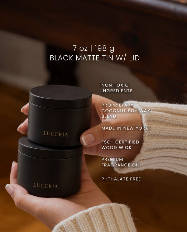 Two black matte cylindrical tin containers with gold 'LUCERIA' branding are stacked in a person's hands. White text on the right lists product details, and the background shows a warm wooden interior.
