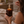 A person in a cozy bedroom setting holds an amber glass 'LUCERIA' candle with a lit wick and flame. White text overlays highlight product features such as '10 oz | 283 g AMBER GLASS CANDLE' and other scent and material claims.