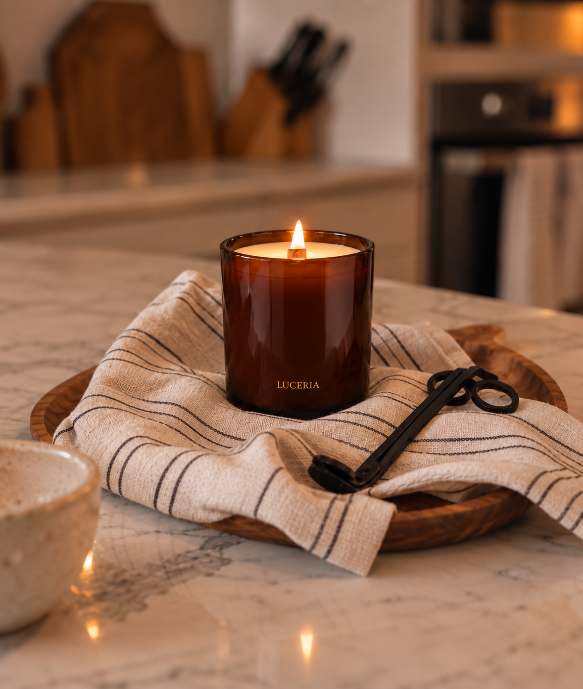 A lit amber glass jar candle with the brand name LUCERIA printed in gold near the base, resting on a striped linen cloth over a wooden tray on a marble countertop in a warm kitchen setting.