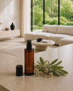 Amber glass Luceria room spray beside fresh sage and chamomile on a light stone surface in a modern minimalist living room.