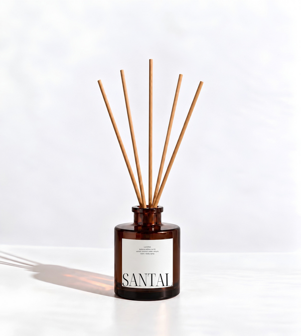A Luceria reed diffuser with a dark amber glass bottle and natural wood reeds, featuring a white label with the brand name 'SANTAL' printed in black. The diffuser is set against a clean, light gray background with a soft shadow cast to the left.