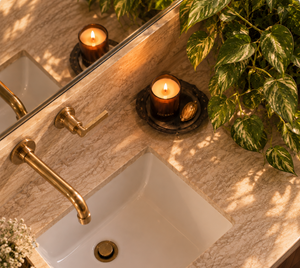 A close-up, high-angle view of a modern bathroom vanity featuring a white rectangular sink, a brushed brass faucet and matching hardware, a lit Luceria candle on a coaster, and a green plant on a marble countertop.