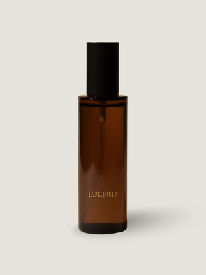 An amber glass bottle with a black cap and gold 'LUCERIA' branding, centered on a light beige background.