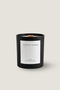 A black glass jar candle with a white label reading 'AUTUMN WOODS' and a small wood wick, centered on a light warm gray background by Luceria Candle Co.