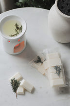 White wax melts with herbs on a marble surface next to a small candle and plant.