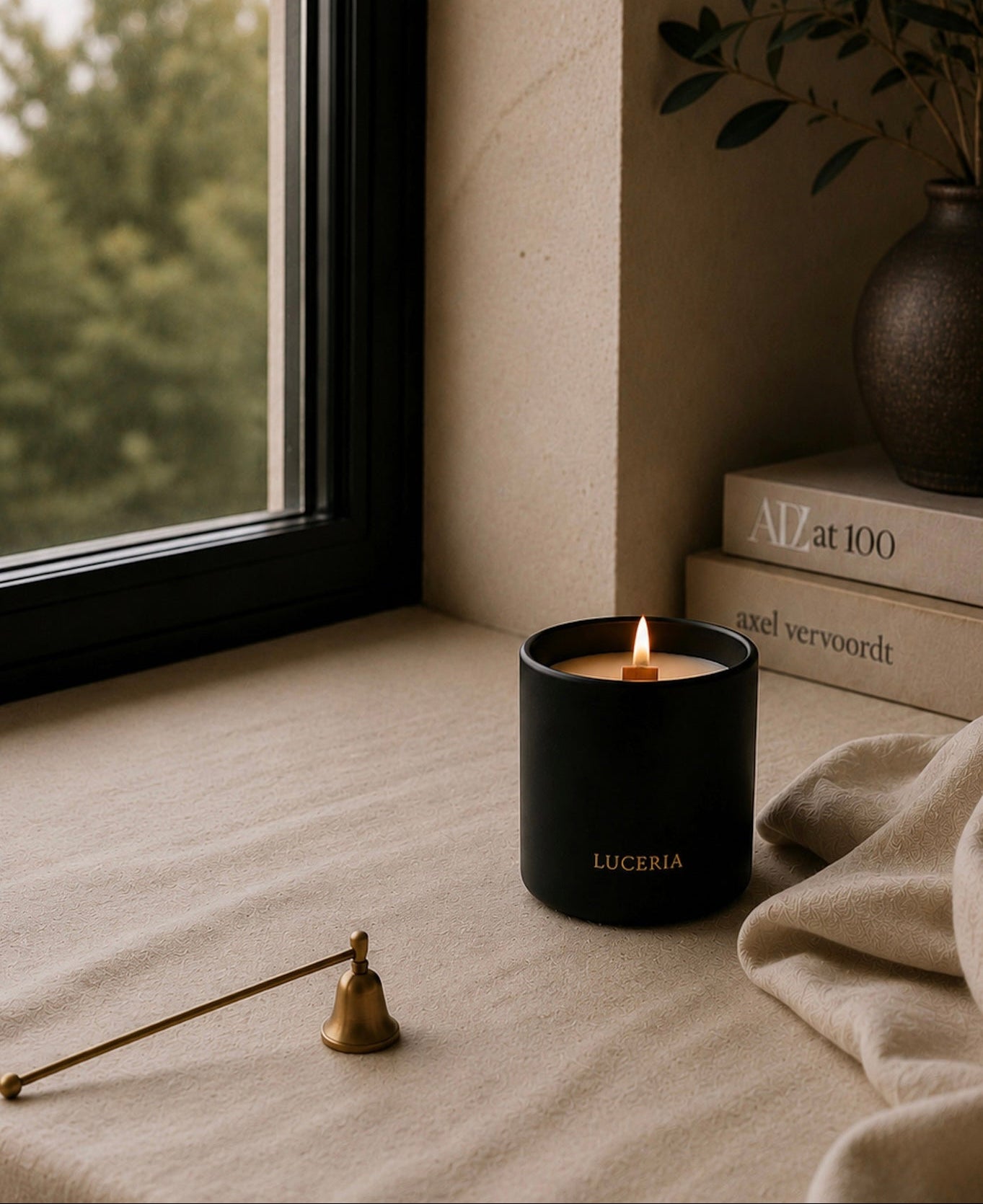 A black matte candle jar with gold 'LUCERIA' branding sits on a beige linen surface by a window. A brass candle snuffer rests nearby, with stacked books and a plant in the background.
