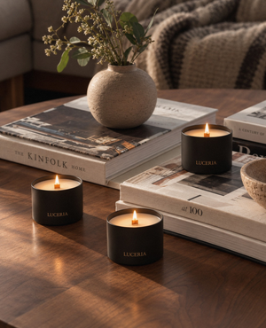 Three black wax candles in matte black jars with gold 'LUCERIA' branding, arranged on a wooden table with stacked books and a small vase in a cozy indoor setting.
