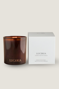 An amber glass candle jar with gold 'LUCERIA' branding stands next to its white square packaging box on a light beige background.