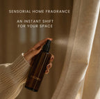A hand in a cream ribbed sweater holds a dark amber spray bottle labeled LUCERIA, spraying a fine mist toward beige curtains. Text at the top reads, 'SENSORIAL HOME FRAGRANCE' and 'AN INSTANT SHIFT FOR YOUR SPACE'.