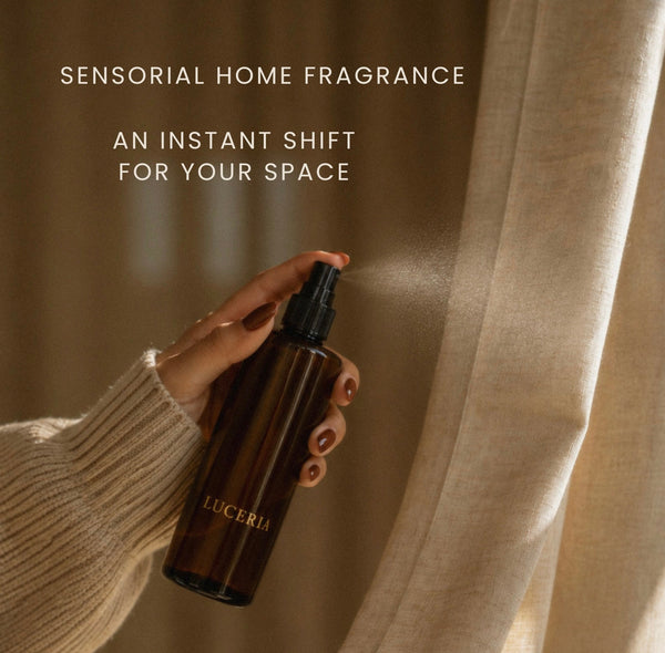 A hand in a cream ribbed sweater holds a dark amber spray bottle labeled LUCERIA, spraying a fine mist toward beige curtains. Text at the top reads, 'SENSORIAL HOME FRAGRANCE' and 'AN INSTANT SHIFT FOR YOUR SPACE'.