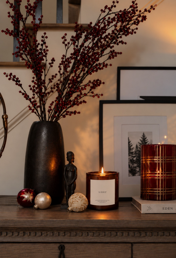 A styled interior tabletop scene with a dark matte vase holding red berry branches, a lit amber Luceria candle with a WINTER label, a red patterned candle holder, ornaments, and framed wall art in a warm room setting.