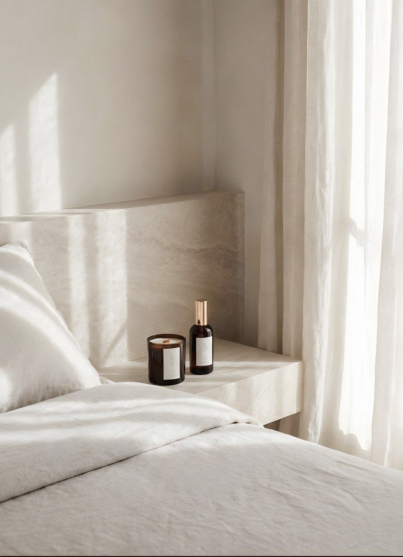 Bedroom with a nightstand holding two Bamber ottles and a Luceria candle, with light filtering through curtains.