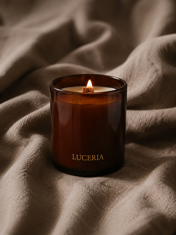 An amber glass candle jar with the brand name LUCERIA printed in gold near the bottom, lit with a small flame. The candle is set against a background of draped beige fabric.