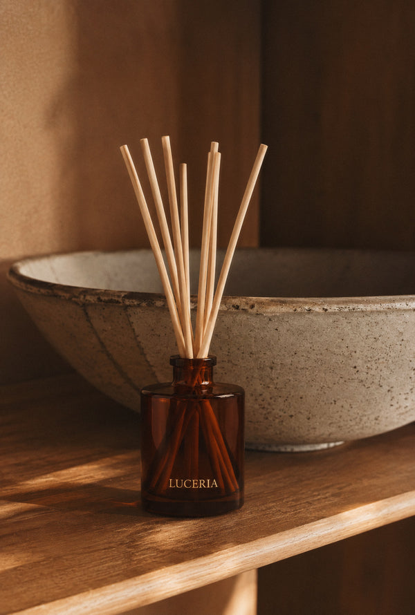 Amber glass reed diffuser bottle with natural wood reeds, labeled LUCERIA, placed on a wooden shelf in front of a speckled ceramic bowl.