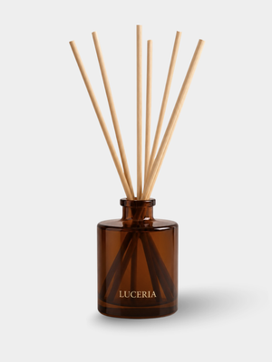 Amber glass reed diffuser bottle with natural wood diffuser sticks, labeled LUCERIA, on a light gray background.