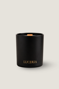 A matte black glass candle jar with gold 'LUCERIA' branding on the front, featuring a natural wood wick, set against a solid light beige background.