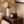 A styled interior scene showing a dark amber glass candle jar labeled LUCERIA next to a matching amber bottle with a gold cap, placed on a wooden side table beside a white ribbed vase of dried flowers.