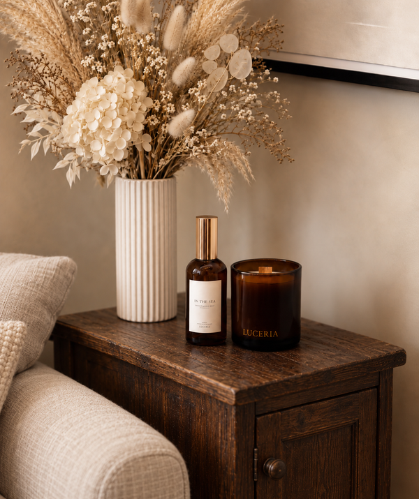 A styled interior scene showing a dark amber glass candle jar labeled LUCERIA next to a matching amber bottle with a gold cap, placed on a wooden side table beside a white ribbed vase of dried flowers.