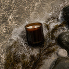 Luceria Sea Salt and Musk candle with a natural wood wick styled with sand, coastal grasses, driftwood, and neutral beach-inspired décor.