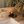 A lit amber glass candle with a black label reading 'LUCERIA' sits on a small black tray on a beige stone bathroom counter next to a white sink and gold faucet, with dried plants reflected in a mirror.