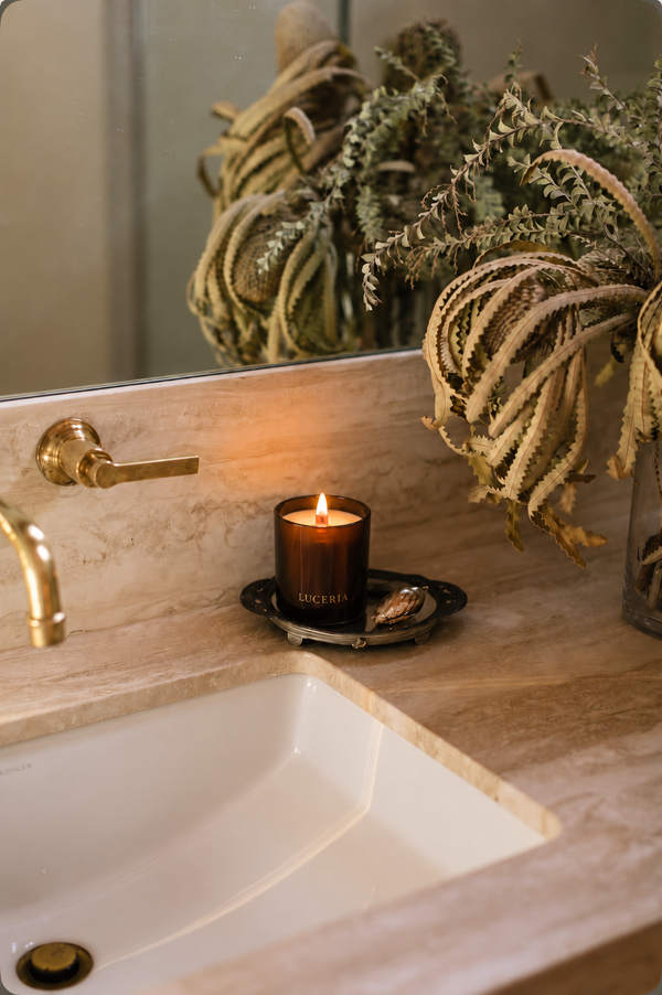 A lit amber glass candle with a black label reading 'LUCERIA' sits on a small black tray on a beige stone bathroom counter next to a white sink and gold faucet, with dried plants reflected in a mirror.