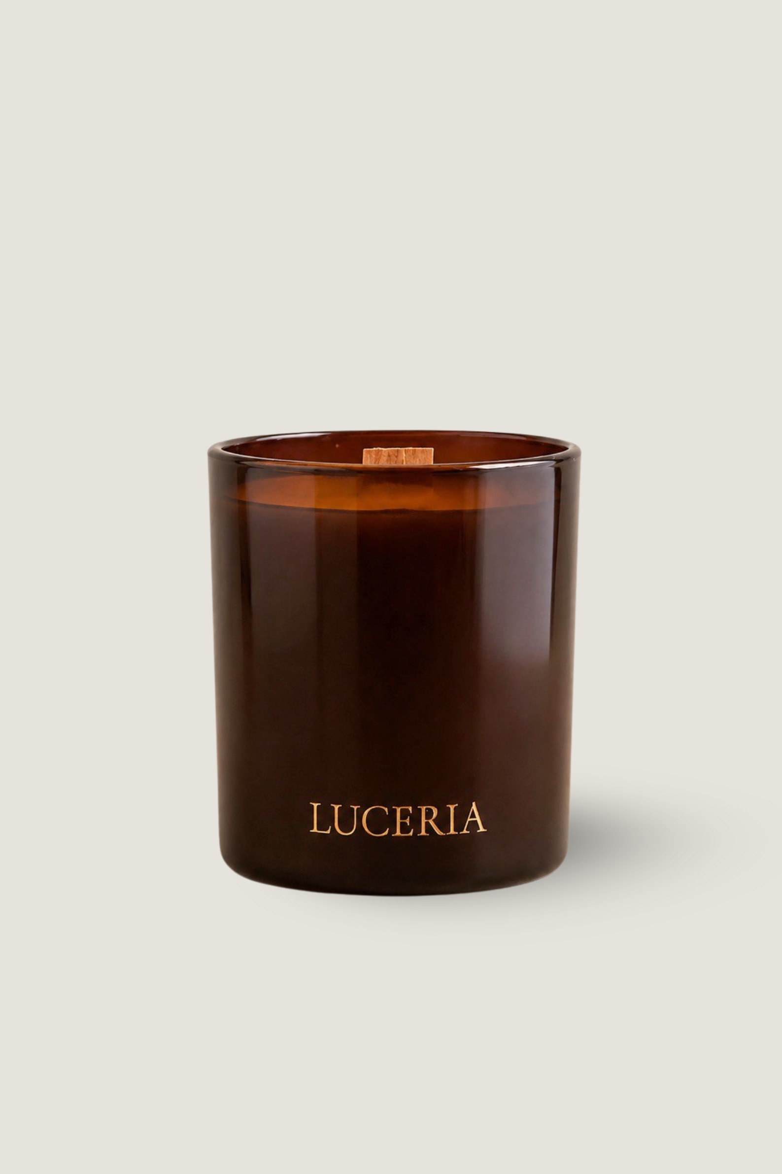An amber glass jar candle with a wick and the brand name 'LUCERIA' printed in gold lettering near the bottom, set against a solid light beige background.