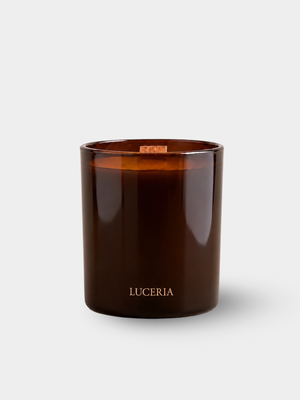An amber glass jar candle with a light wood wick and the brand name 'LUCERIA' printed in gold near the bottom, set against a clean light gray background.