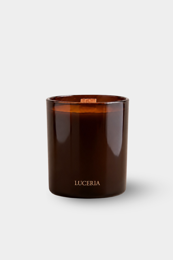 An amber glass jar candle with a light wood wick and the brand name 'LUCERIA' printed in gold near the bottom, set against a clean light gray background.