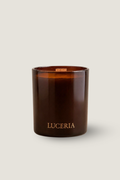 An amber glass jar candle with a wick and the brand name 'LUCERIA' printed in gold at the bottom, set against a solid light beige background.
