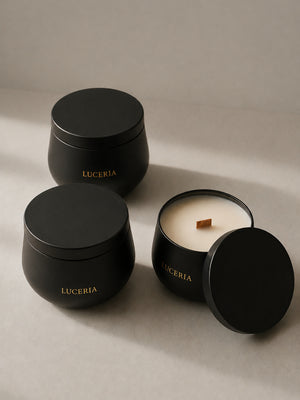Three matte black candle containers from Luceria on a warm beige surface, with one open candle showing white wax and a wooden wick alongside two closed jars.