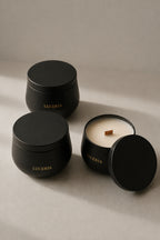 Three matte black candle containers from Luceria on a warm beige surface, with one open candle showing white wax and a wooden wick alongside two closed jars.