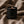 A black matte candle jar with the brand name 'LUCERIA' in gold lettering, lit with a small flame, resting on a draped brown fabric background.