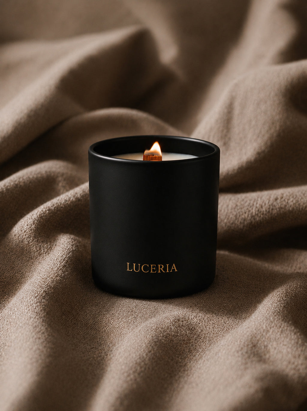 A black matte candle jar with the brand name 'LUCERIA' in gold lettering, lit with a small flame, resting on a draped brown fabric background.