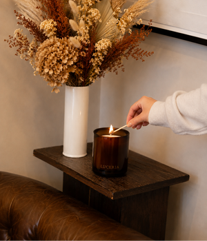 A person's hand lighting a dark amber glass 'LUCERIA' candle on a wooden side table, with a white vase of dried flowers in the background.
