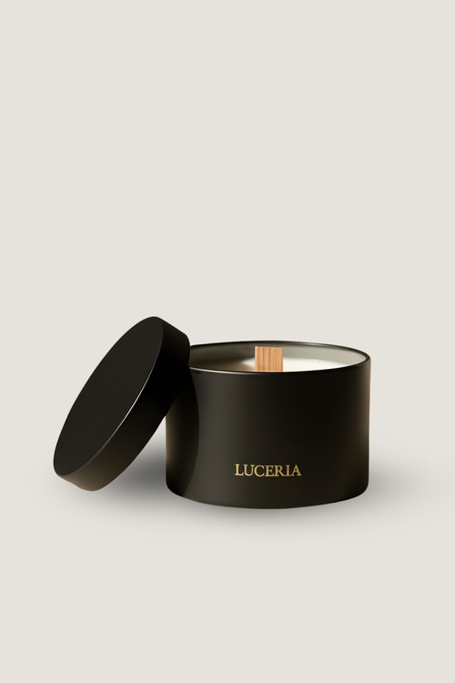 A black cylindrical candle jar with its lid leaning against it on a light warm-gray background. The jar is labeled 'LUCERIA' in gold lettering, and a small wooden wick is visible inside.