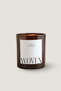 A Luceria amber glass jar candle with a white rectangular label reading 'WOVEN' in black serif letters, shown on a plain light beige background.