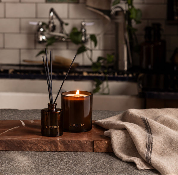 A lit amber glass candle and matching amber reed diffuser bottle with reeds, both branded LUCERIA, sit on a wooden board on a countertop in a real bathroom setting with a towel nearby.