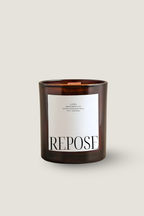 A Luceria amber glass jar candle with a white rectangular label reading REPOSE, photographed on a plain light beige background.