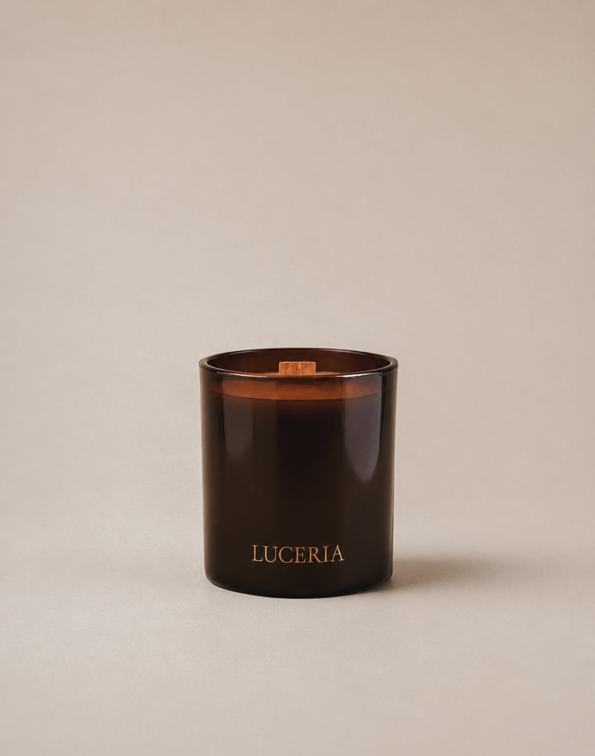 A amber glass coconut soy wax candle with wood wick labeled ‘LUCERIA’ with gold lettering on a neutral beige backdrop with soft diffused lighting. 