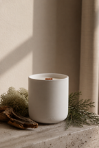 White ceramic Luceria candle scented with oakmoss and white cedar, featuring a natural wood wick on a stone surface with moss and driftwood.