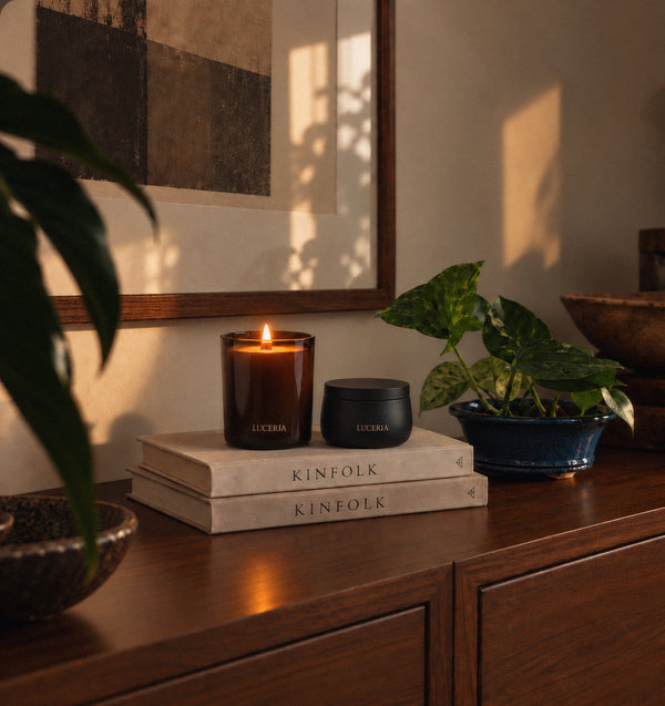 A lit amber glass candle and a small matte black jar both labeled Luceria sit on stacked Kinfolk books on a wooden console, with a potted plant and warm sunlight in a styled interior setting.