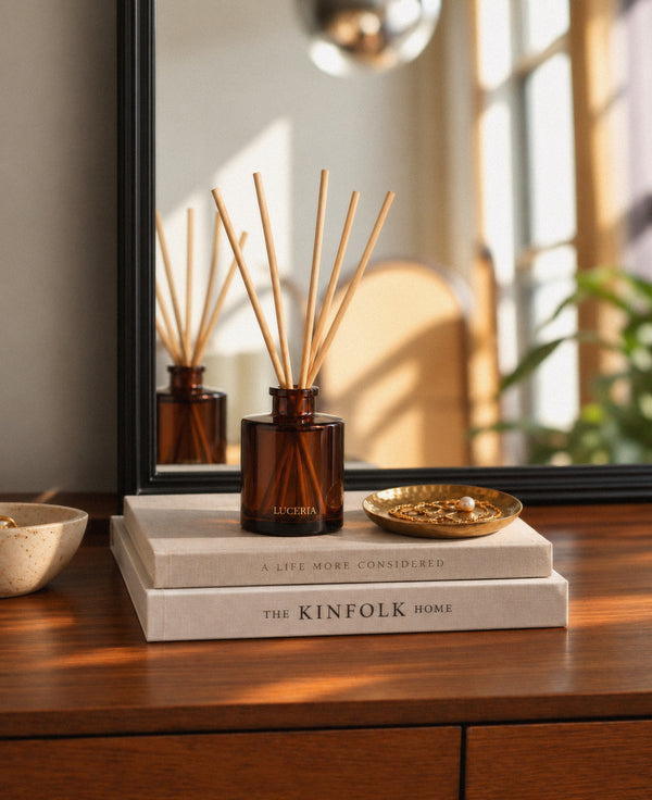 Amber glass Luceria reed diffuser bottle with wooden reeds, styled on stacked books on a wooden table with a mirror and warm window light in the background.