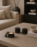 Three black cylindrical candles from the brand Luceria are arranged on a beige stone coffee table in a modern, neutral-toned living room. One candle is open, showing a lit candle inside, while the other two are closed with gold lettering.