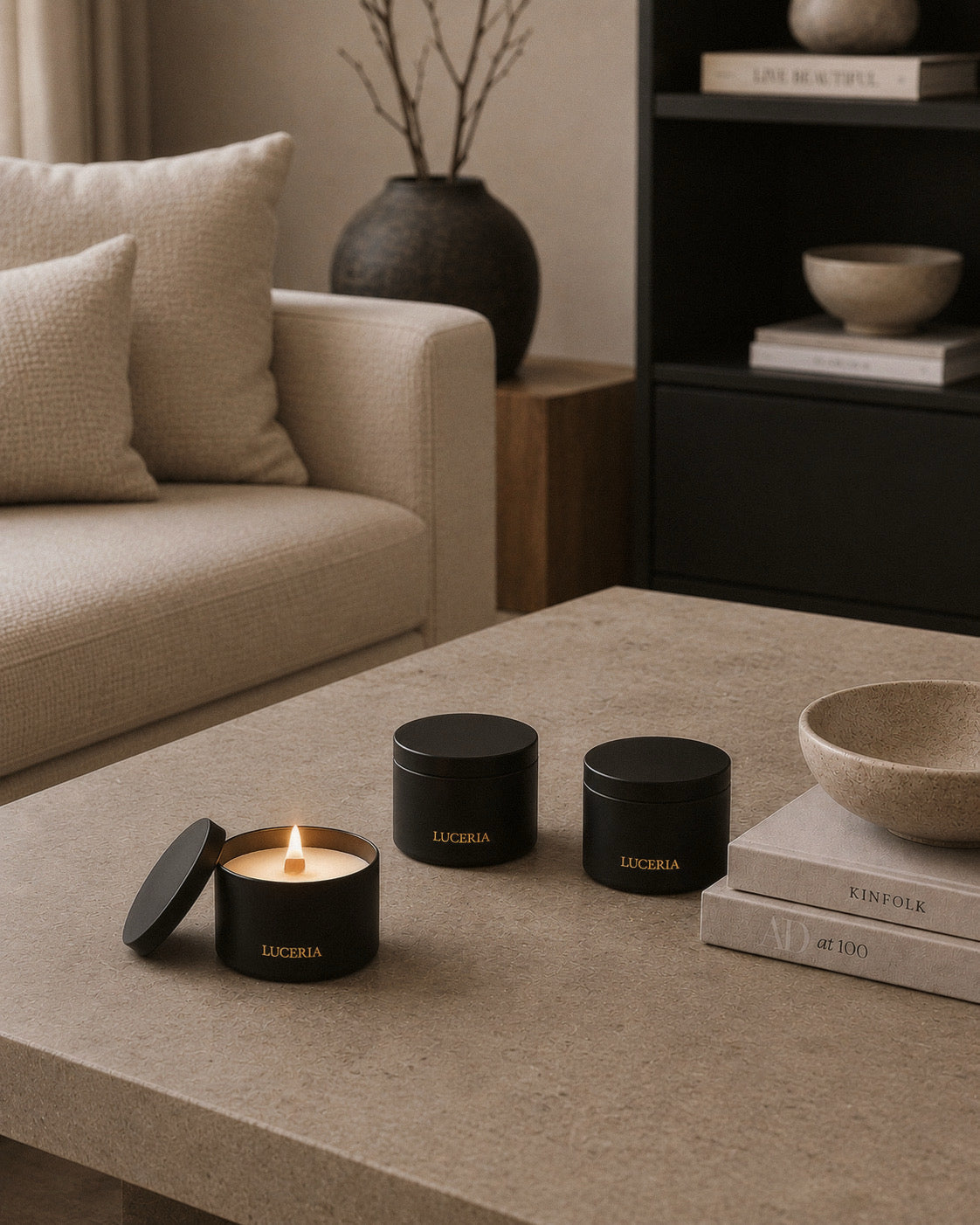 Three black cylindrical candles from the brand Luceria are arranged on a beige stone coffee table in a modern, neutral-toned living room. One candle is open, showing a lit candle inside, while the other two are closed with gold lettering.