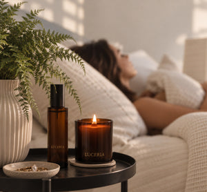 A styled bedroom scene with a woman lying on a bed in the background, while a black side table in the foreground holds a lit amber glass candle and a matching dark bottle labeled LUCERIA, next to a potted fern in a ribbed white vase.